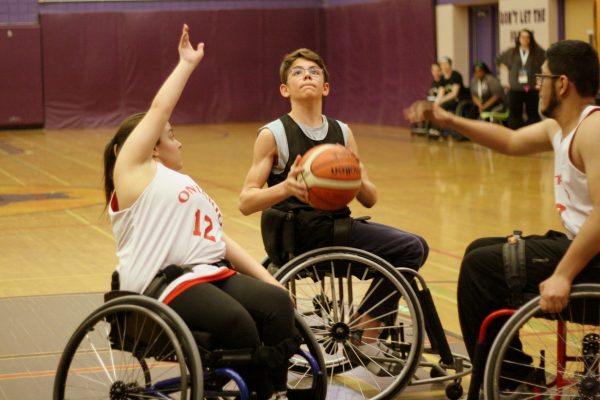 A young boy in a wheelchair prepares to shoot a basketball while two athletes, a boy and a girl, raise their hands to defend the shot