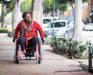 A Black woman wheels along the sidewalk in a pink wheelchair. She is smiling and wearing a red jacket, black pants, black gloves, and a multi-coloured knit scarf